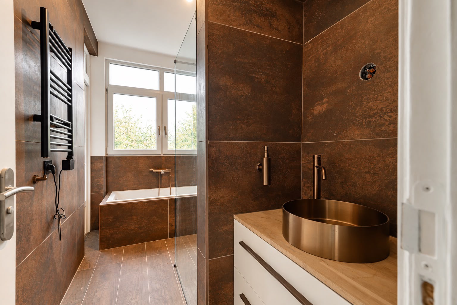 Overzicht van de gerenoveerde badkamer met bad, inloopdouche, wastafelmeubel en bruine wandtegels.